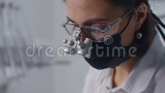 一位年轻的女牙医戴着双筒望远镜和一间白色办公室正在检查和治疗一位女性病人。 牙科治疗和视频