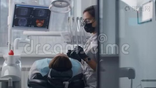 一位年轻的女牙医戴着双筒望远镜和一件白色外套开始检查和治疗一位女性病人。 牙科治疗视频