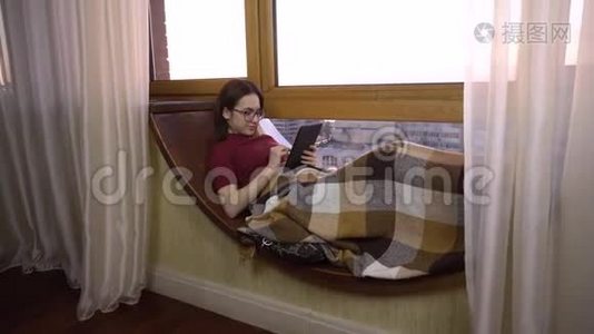 一位年轻女子正在平板电脑上发短信。 一个女孩躺在窗边的窗台上，手里拿着一块平板电脑。 外部视频
