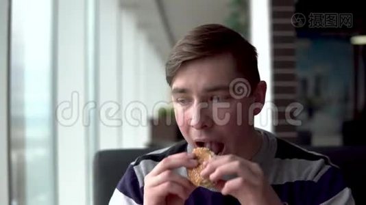 一个年轻人正在吃汉堡。 一个男人坐在全景窗旁的咖啡馆里，吃快餐。视频