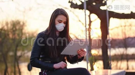 有魅力的女人独自在公园里坐下来，她戴着防护面罩在平板电脑上工作，以确保安全视频