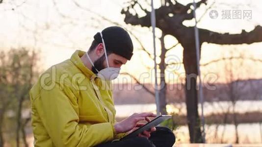 在公园里，日落时分，一个男人坐在椅子上时用电子平板电脑做特写，他戴着一个防护面具视频