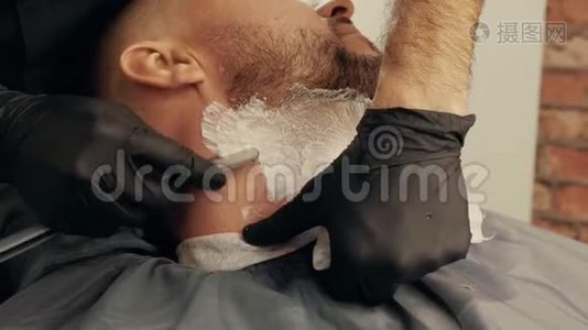 在男式沙龙里用直剃须刀把理发师的手刮胡子。 用剃须刀和泡沫刮胡子视频