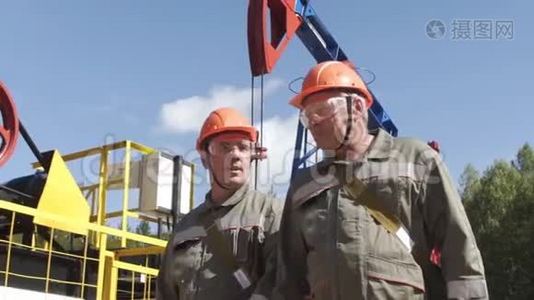 两名戴着橙色头盔的石油工人在油泵千斤顶附近行走和交谈。 石油工程师监督原油现场视频