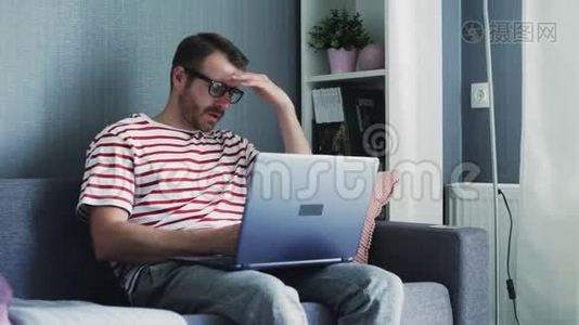 疲劳的年轻人戴着眼镜，坐在家里的沙发上，在笔记本电脑上工作。视频