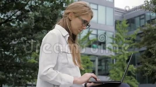 年轻的商务女性在绿色公园的办公楼旁使用便携式电脑。视频