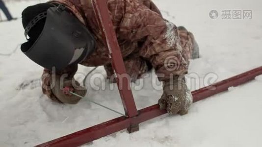 一名焊工在雪中跪着焊接金属框架的特写。 穿着冬装、手套和电焊面罩的人视频