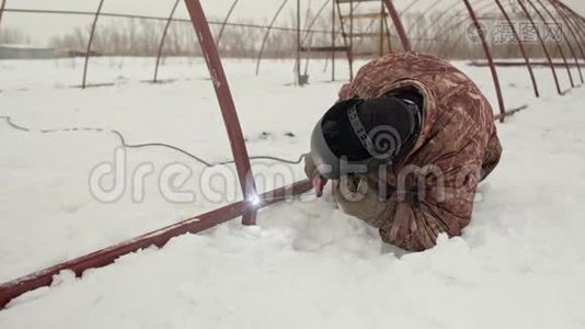 一个穿着温暖的冬装和焊接面罩的人正在地面上焊接一个金属结构。 专业焊工焊接视频