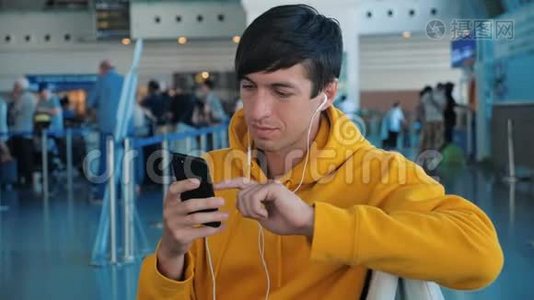 年轻的男性，穿着黄色连帽衫，在机场等候飞机，使用带耳机的智能手机视频