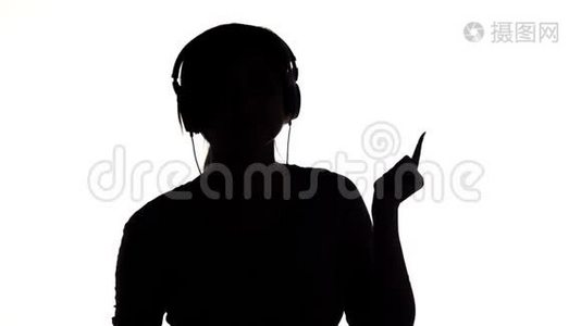 一个年轻快乐的女人戴着大耳机听音乐，拿着智能手机的剪影，女孩在白色的隔离巴上跳舞视频