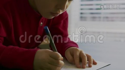 一个穿着红色毛衣的小男孩坐在桌子旁，手里拿着一支钢笔，在空白的白色上写字或画画视频