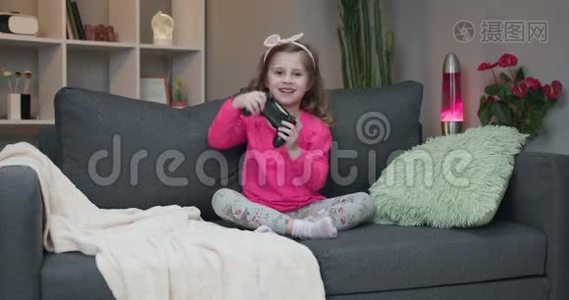 年轻的小女孩坐在沙发上在家里的客厅里播放视频。 兴奋的玩家女孩手拿操纵杆视频