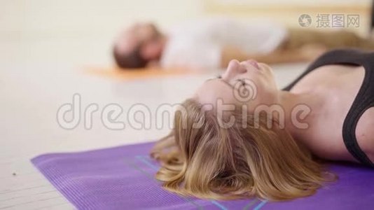 侧视运动的女人和男人一起练习瑜伽，躺在白色瑜伽工作室的休息姿势萨瓦萨纳。视频