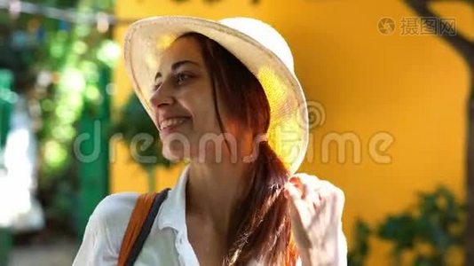 一位戴着草帽、白衬衫、微笑着对着镜头的美丽旅游妇女站在明亮的橙色墙上视频