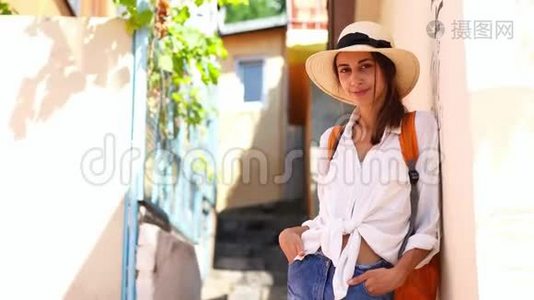年轻优雅的女游客，穿着草帽、牛仔裤短裤、白衬衫和橙色背包，在狭窄的街道上散步视频
