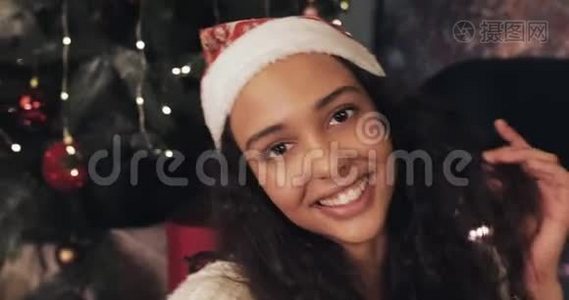 圣诞老人帽子里迷人的年轻微笑女子在智能手机上自拍，坐在圣诞树附近的家中视频