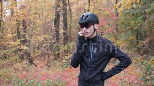 年轻的职业男性自行车运动员在秋季公园骑自行车锻炼前打电话。 紧张的骑自行车的家伙说视频