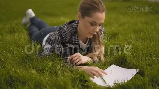热情的女艺术家躺在草地上，在笔记本上画画。 公园，绿草如茵.. 4K视频