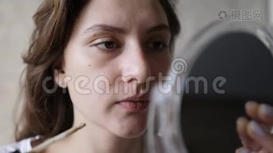 一个年轻的女人在她的眼睛下面用刷子涂抹遮瑕膏，然后看着一面小镜子。 基本化妆视频