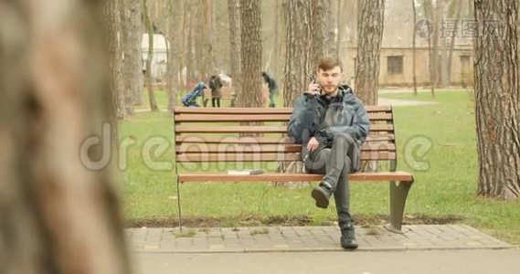 年轻的胡须男拨打电话，开始在电话上交谈，微笑着，享受着秋公园坐在长凳上的谈话。视频