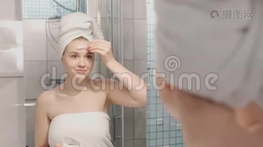 一张美丽的年轻女子的肖像，穿着浴袍和毛巾站在浴室里，在浴室下面敲击补片视频