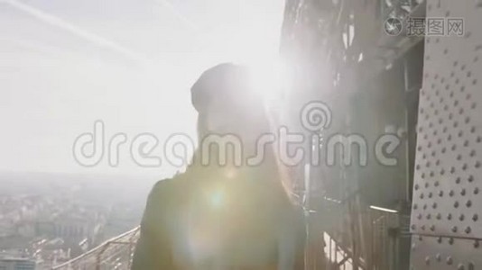 惊人的镜头耀斑拍摄，年轻的旅游妇女走向相机微笑沿着埃菲尔铁塔顶部缓慢的运动。视频