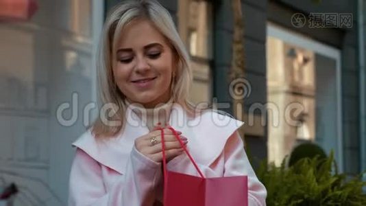 一张年轻漂亮的金发女郎穿着粉红色外套，站在街上，拿着一个红色的纸袋，微笑着的肖像。视频