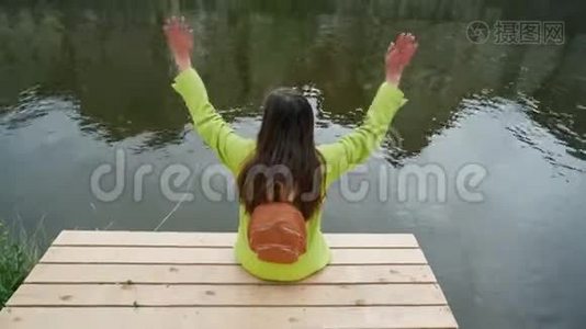 穿着黄色夹克的长发女人，背包坐在河边或池塘的木墩上，挥舞着双手。 传统视频