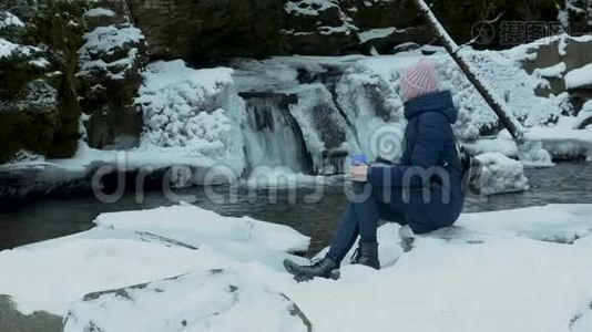 穿着蓝色外套的女孩坐在一片冰雪覆盖的针叶林中，脸上挂着一个杯子，背景是冰冷的瀑布和岩石。 冬季视频