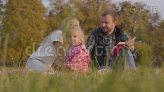 幸福的一家人坐在秋天的草地上，吃苹果，读红色封面的书。 有胡子的白种人视频