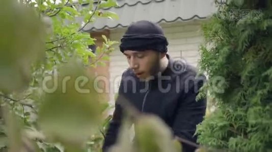 一个穿着黑色衣服的年轻白人小偷闯入家门前藏在树下的画像。那家伙要视频