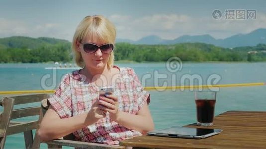 商务女性在度假时使用智能手机。 坐在湖边的一家咖啡馆里的一张桌子上，背景是群山。视频