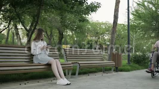 在公园里坐轮椅的年轻人和坐在长凳上看着他视频
