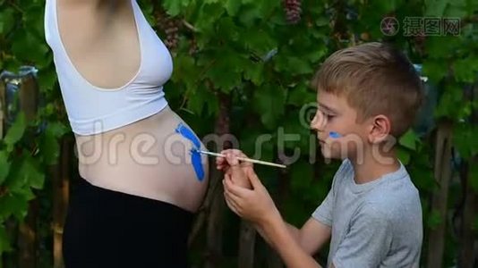 笑的男孩用刷子在他怀孕妈妈宝宝的肚子上画`脚印。 怀孕的概念。 特写视频