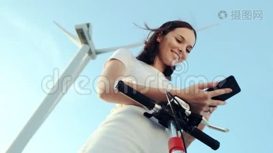 这个女孩在她的电动滑板车旁边使用一部智能手机，背景是风力发电机。视频