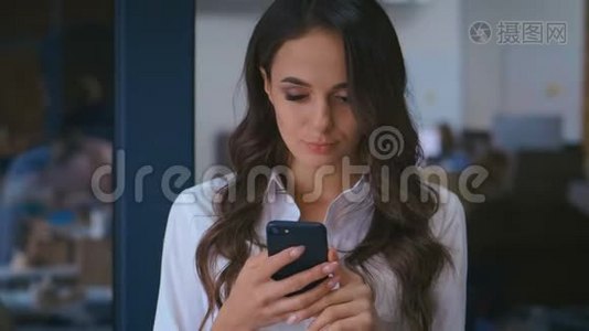办公室使用智能手机的美丽年轻女性肖像。 穿白色衬衫的商务女士在她的手机上输入信息视频