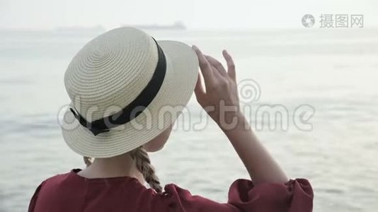 日落时分，一个穿着夏红色连衣裙和草帽的白人女孩坐在海滨的近景中视频