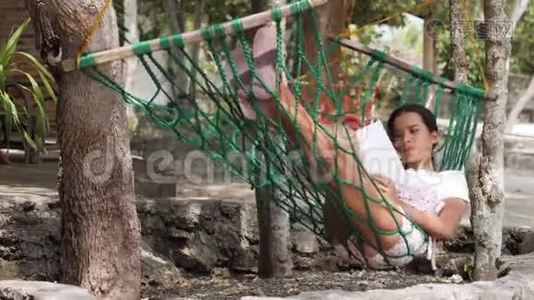 一个年轻的女人在户外树下的一个旋转吊床上看书。视频