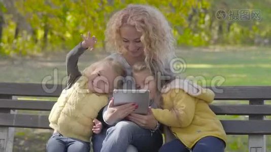 年轻的白种人，带着一头金色卷发，坐在秋天公园的长凳上，和两个漂亮的小女孩在一起视频