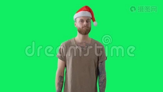身穿绿色t恤，戴着红色圣诞帽，长着胡须的年轻纹身男正在彩色钥匙背景上接受礼物视频