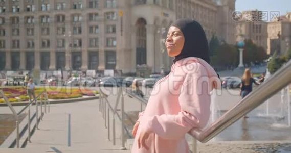 年轻迷人的非洲裔美国穆斯林女孩戴着头巾站在喷泉旁开心地笑着的特写照片视频