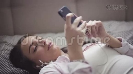 年轻女子躺在床上，手里拿着智能手机，耳机插上，女孩听音乐或播客。视频