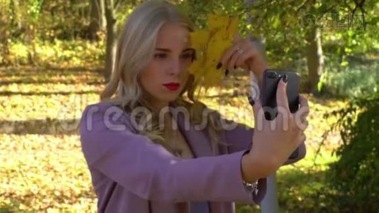 秋叶年轻迷人的金发女郎在秋林公园的手机上自拍。视频