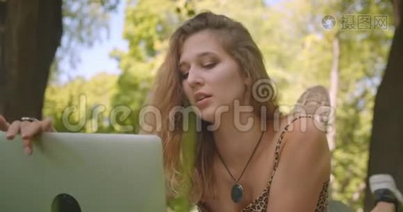 一位年轻漂亮的白种人女性的特写照片，她在笔记本电脑上打着视频，开心地躺在草地上的地毯上视频