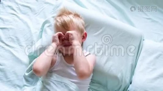 一个金发小男孩躺在床上微笑，这个男孩在清晨用双手揉眼睛。 是时候醒来了视频