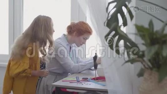 美丽的红发女人缝衣服坐在桌子旁边的窗户在灯光室。 小女儿看上去视频