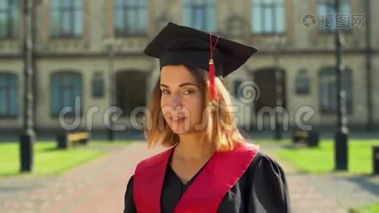 戴着黑色斗篷和帽子的漂亮女孩站在大学大楼前看镜头的肖像。 青年视频