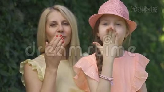 金发母亲和她的小女儿坐在夏天的公园里，一边看着镜头，一边送空气亲吻的肖像视频
