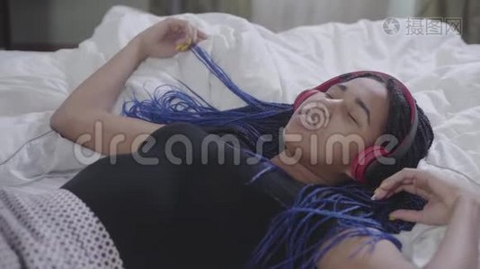可爱的非洲裔美国女孩戴着耳机躺在白色床上的肖像，眼睛紧闭，头部移动。 年轻女子视频