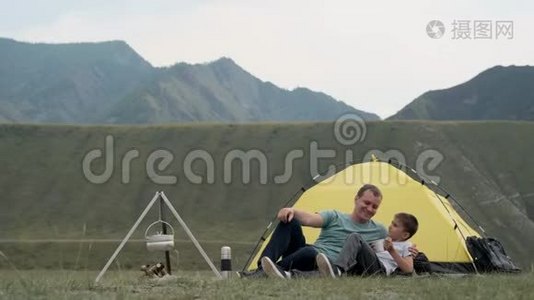 快乐的父子一起放松，在群山的背景下，在帐篷前欢乐。视频